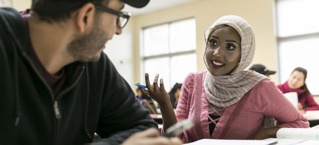 English as a second language (ESL) class at the Eastview Campus. A student in a headscarf and pink blouse talks with a student wearing glasses and a ballcap.