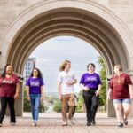 A group of smiling students walk under an archway on a university campus.
