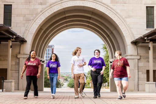 A group of smiling students walk under an archway on a university campus.