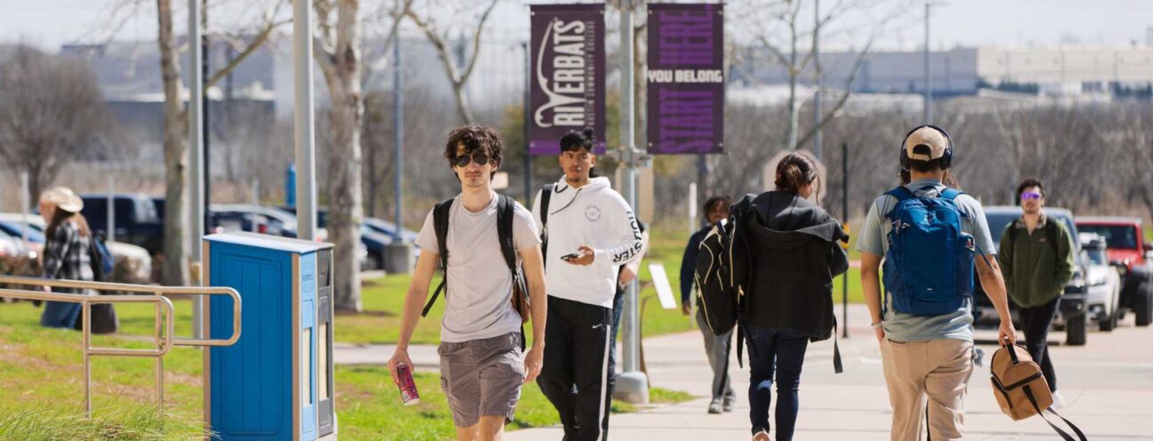 ACC students walk along the sidewalk near a campus building, engaged in conversation and carrying backpacks.