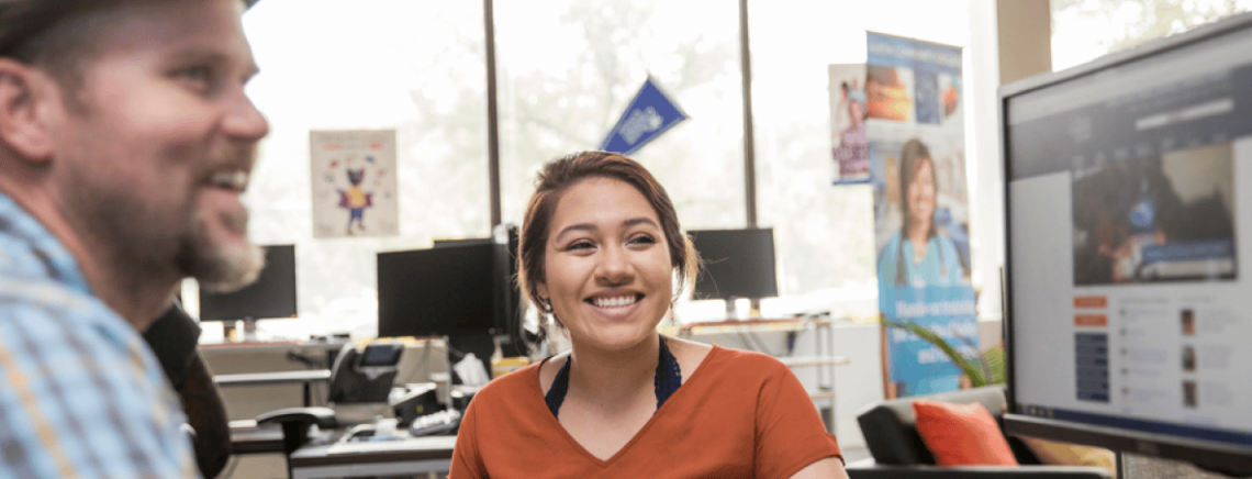 Two people smiling and discussing something in a modern office setting with computers and a window in the background. A woman in the foreground wears an orange shirt, and a man in a plaid shirt and hat is slightly out of focus. A banner for Austin Community College is visible in the background.