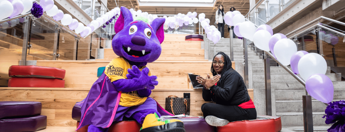 A person poses with RB the Riverbat from Austin Community College on a set of indoor stairs decorated with purple and white balloons. The mascot is dressed in a cape and yellow t-shirt with the college logo, and the person next to the mascot is seated, smiling, and making a hand gesture. The setting appears to be a modern, festive event.