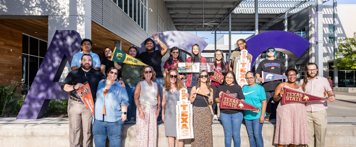 A diverse group of students and staff stands in front of a large purple "ACC" sign, representing the college. They are holding various school banners and signs, showcasing their affiliations with different institutions. The group is smiling and posing together in a sunny outdoor setting, with modern architecture in the background.