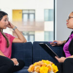 A young woman in a pink top and black pants speaks with an older woman in black blazer and purple blouse, and black pants. They sit on a couch in front of a window with a plant in the background. The older woman holds a clipboard and listens intently to the young woman.