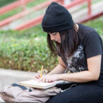 A young woman wearing a black beanie and a graphic t-shirt sits on a stone bench on the ACC Riverside campus. She is focused on writing in a notebook, with a backpack beside her. In the background, there are green trees and a pathway with red railings. The scene conveys a calm and studious atmosphere.