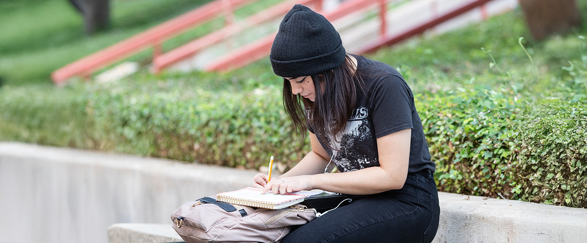 A young woman wearing a black beanie and a graphic t-shirt sits on a stone bench on the ACC Riverside campus. She is focused on writing in a notebook, with a backpack beside her. In the background, there are green trees and a pathway with red railings. The scene conveys a calm and studious atmosphere.