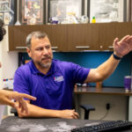 A male student with black hair, brown skin, and a white shirt sits with a male ACC advisor in a purple ACC polo shirt choosing classes in New Student Orientation.