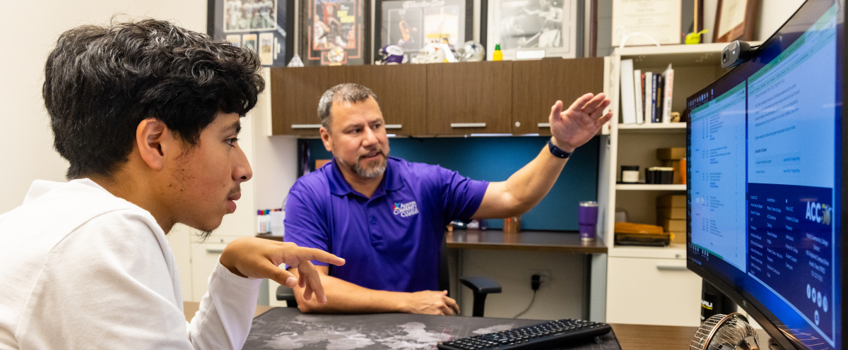 A male student with black hair, brown skin, and a white shirt sits with a male ACC advisor in a purple ACC polo shirt choosing classes in New Student Orientation.