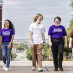 Five students wearing Texas State and ACC Riverbats T shirts walk under the arch on the Texas State campus after successfully transferring through the Bats to Cats program.