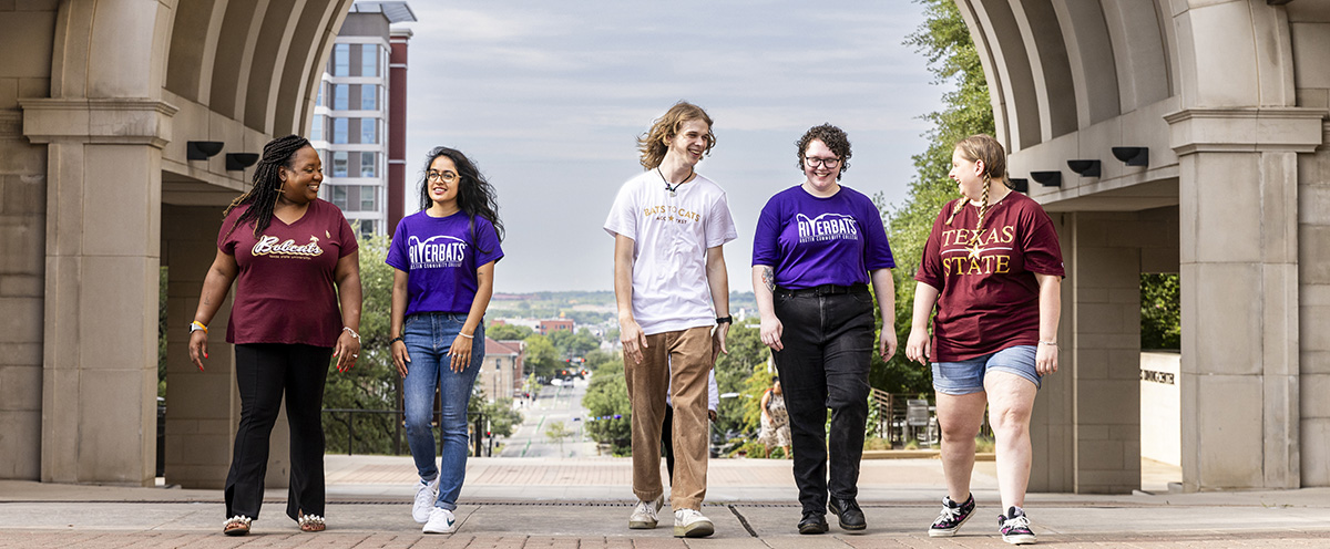 Five students wearing Texas State and ACC Riverbats T shirts walk under the arch on the Texas State campus after successfully transferring through the Bats to Cats program.